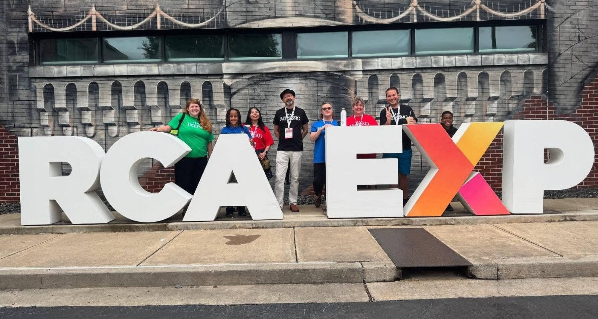 A group of nine people, including children and adults, pose behind large white letters spelling 'RCA EXP' on a city sidewalk